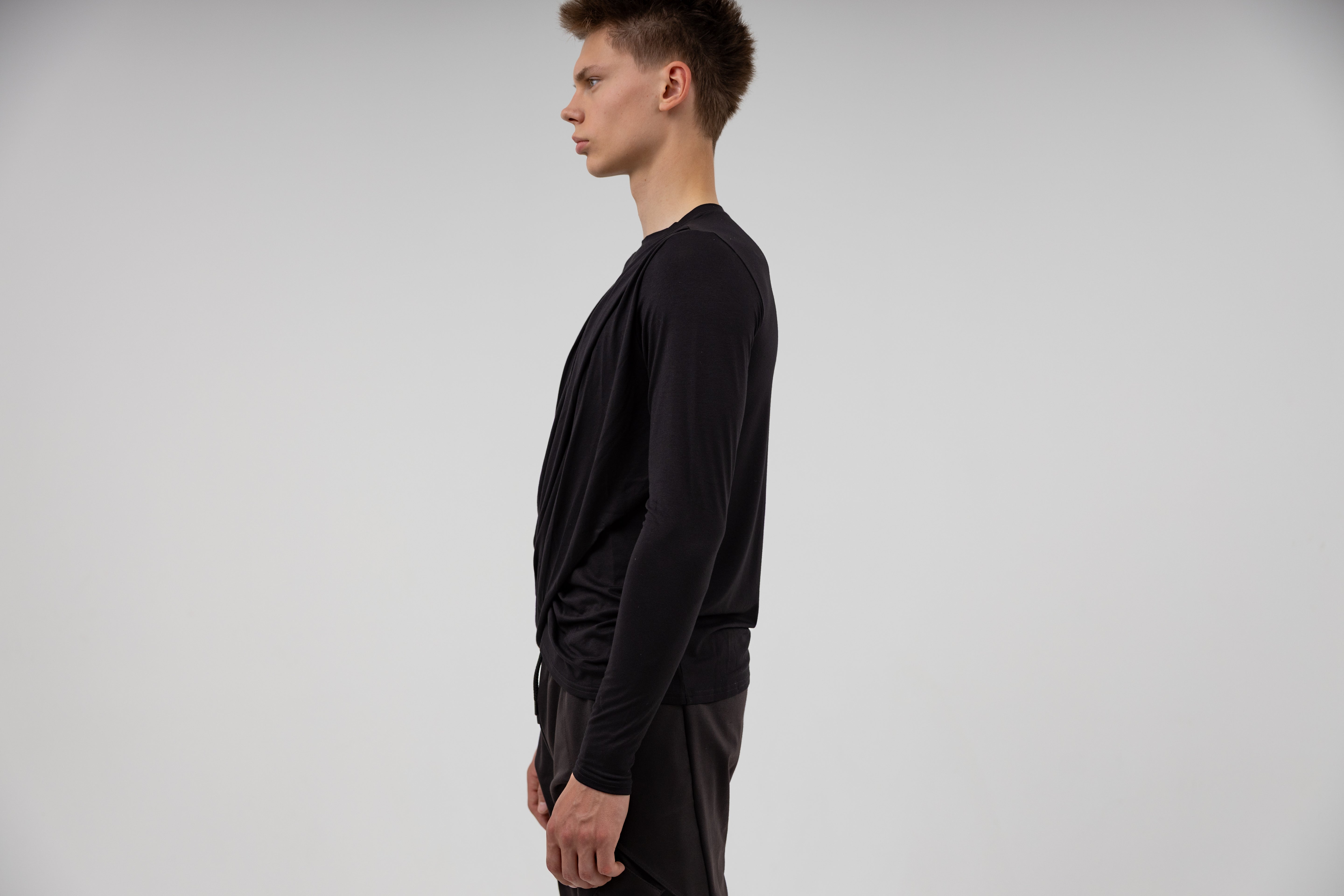 sideview of a male model wearing black long-sleeve shirt and dark urban streetwear cargo pants on a light gray background