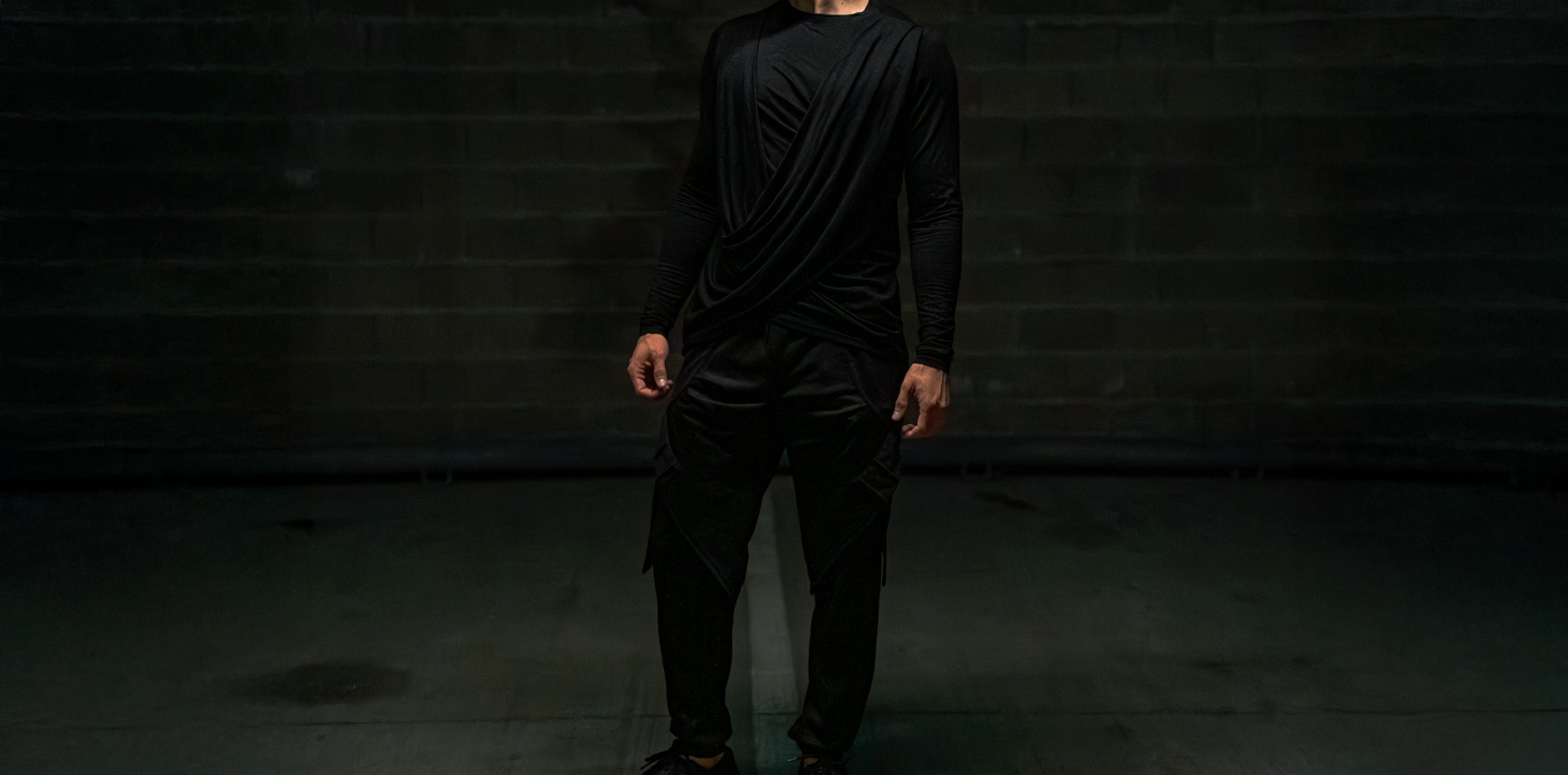 new trending dark black urban casual smart streetwear outfit with male person standing against a dark brick wall.
