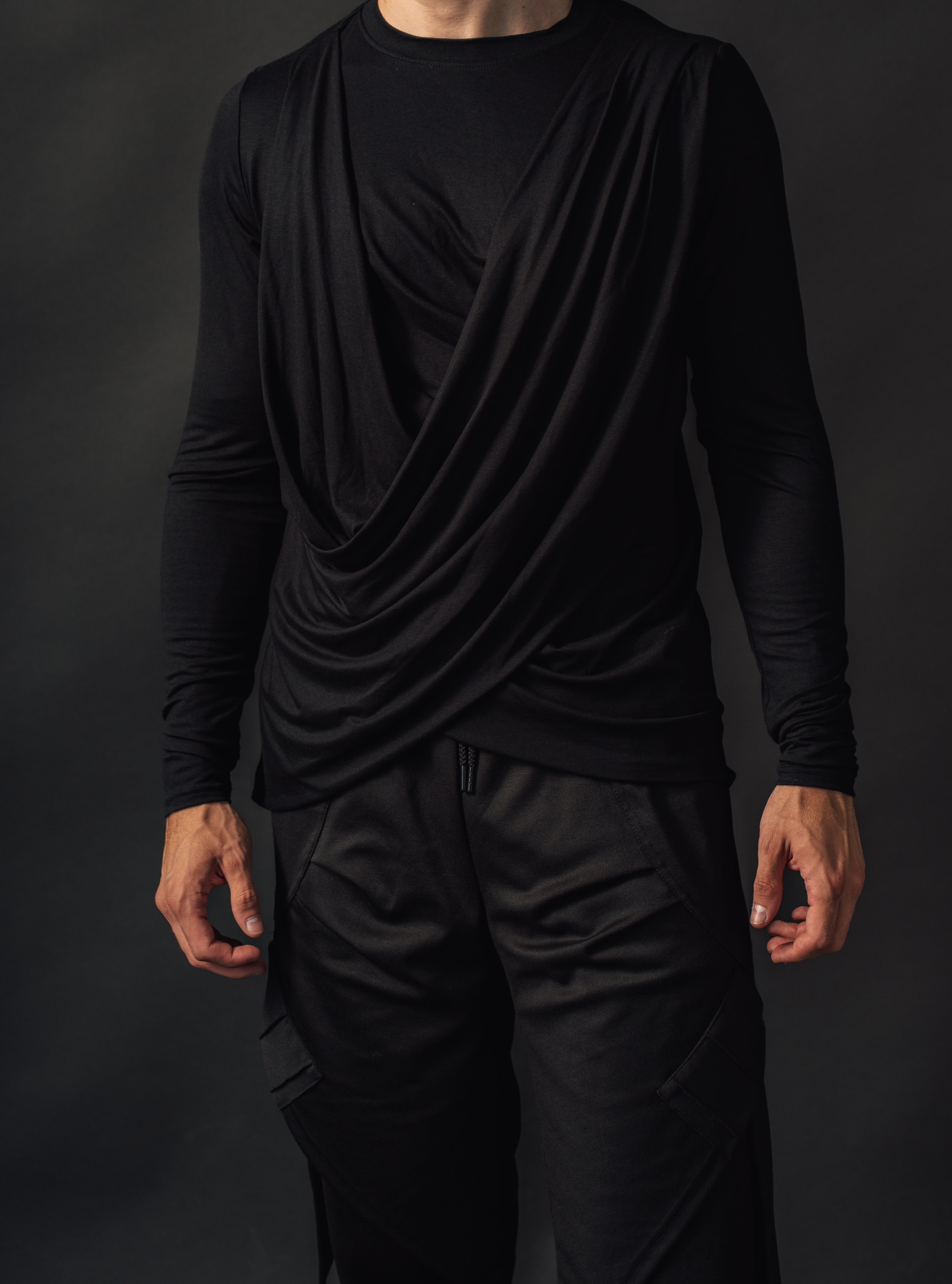 Male person wearing a black draped fashion aesthetic top and pants against a dark background