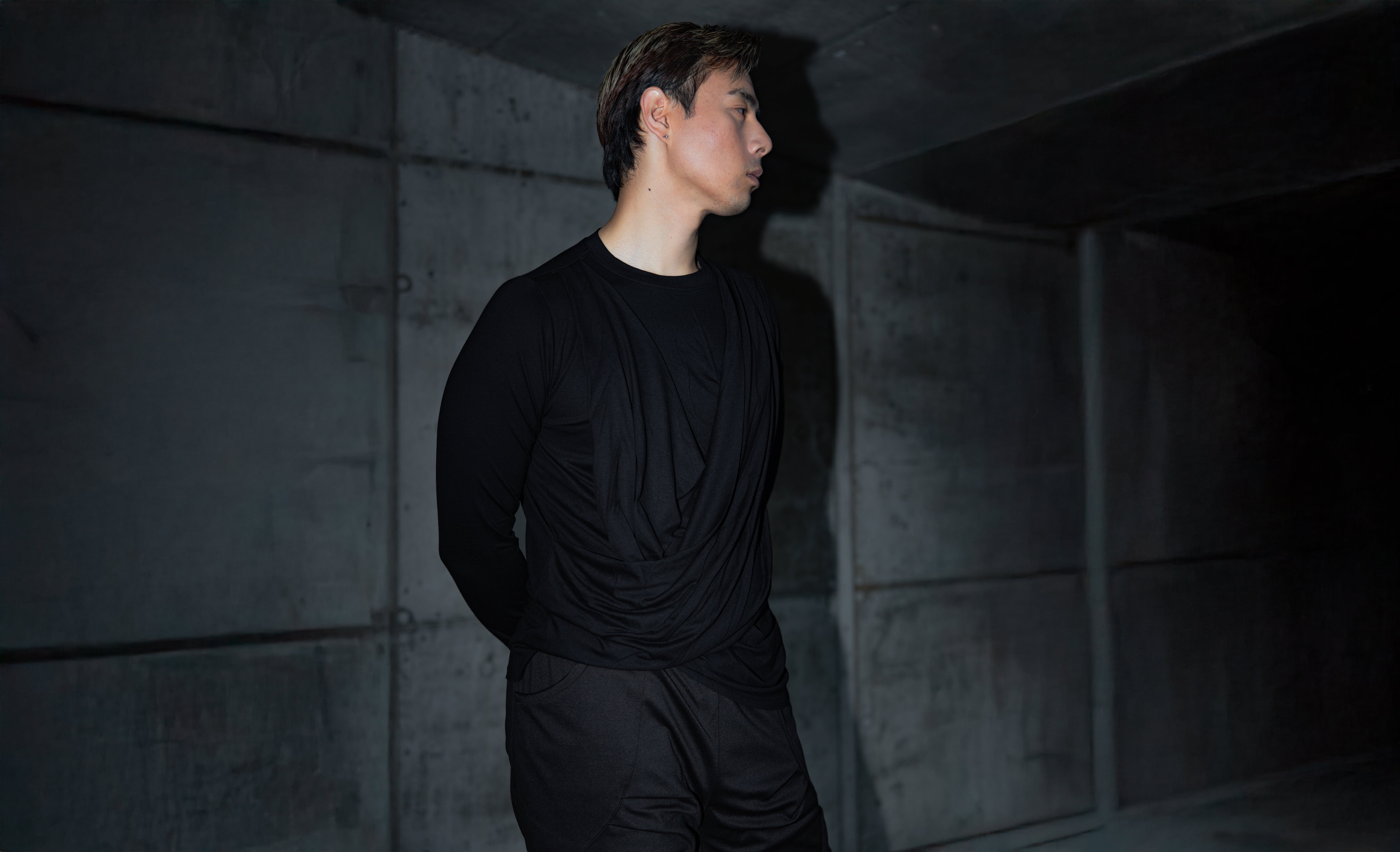Person wearing a black dark fashion clothing outfit standing in a dark, industrial setting