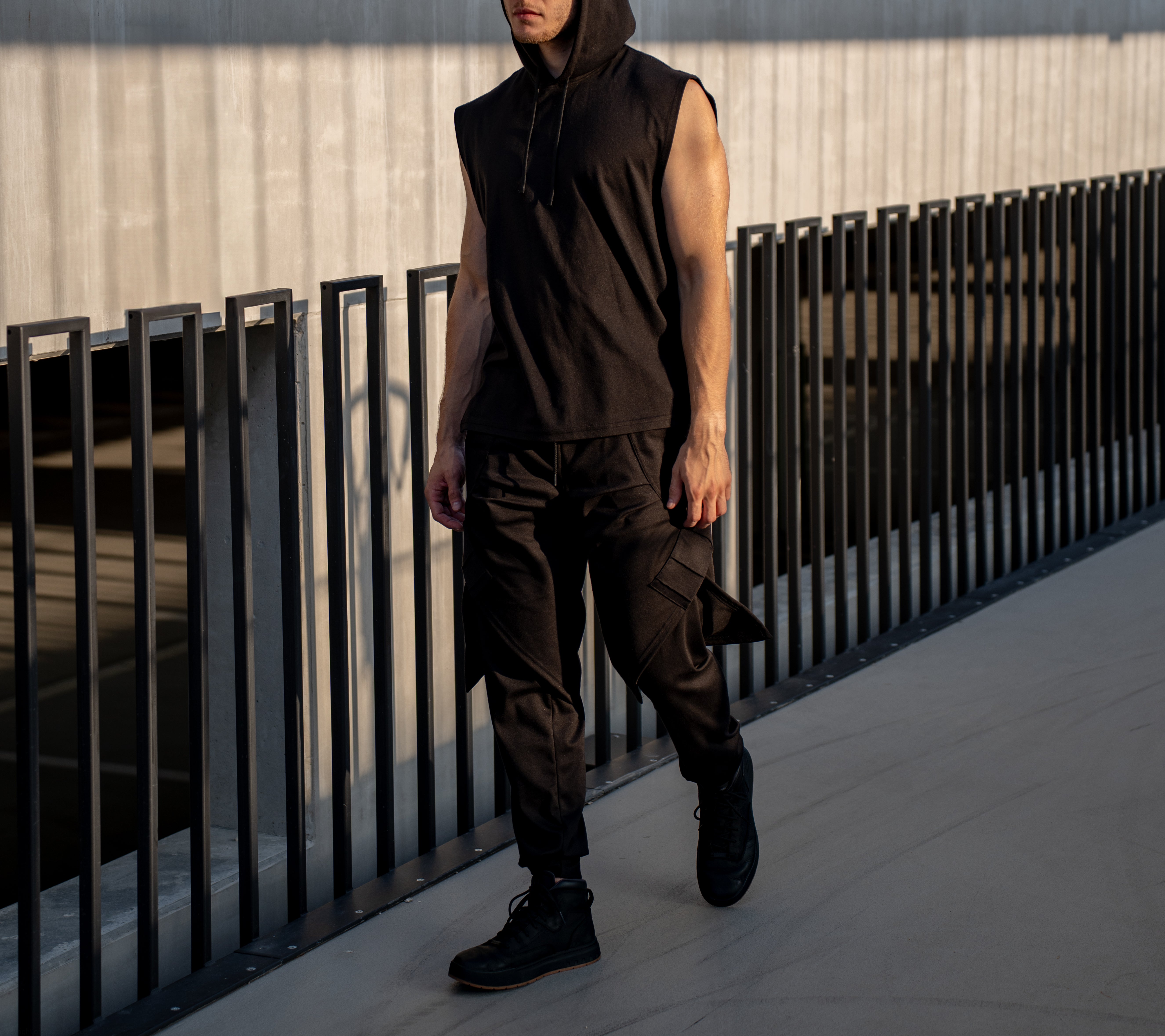 Person wearing a black sleeveless hoodie and layered fashion trending pants standing on a rooftop with a metal railing.