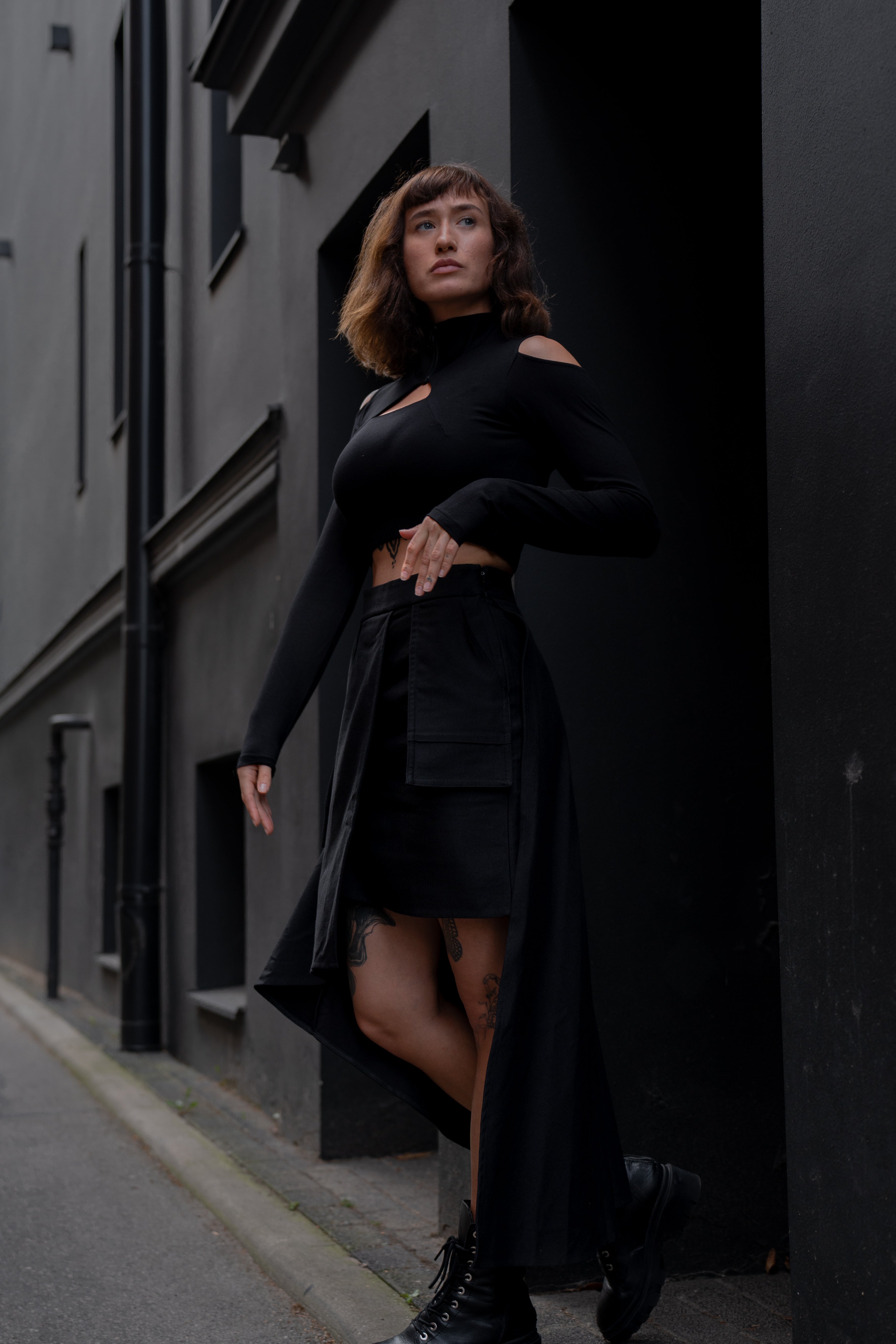 Woman in a black outfit standing against a dark building wall.