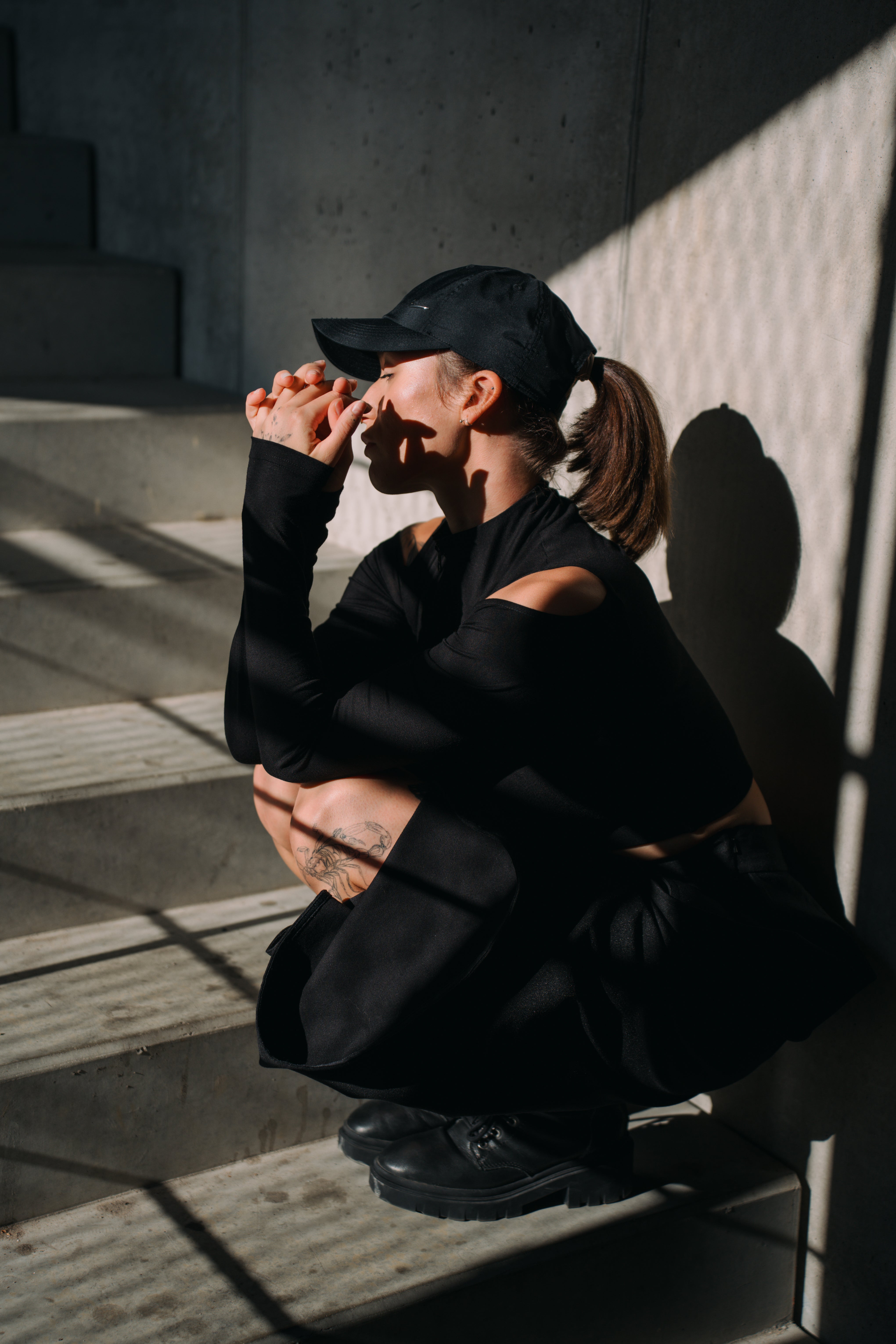 Person in black outfit squatting on a concrete surface with shadows cast by sunlight. 