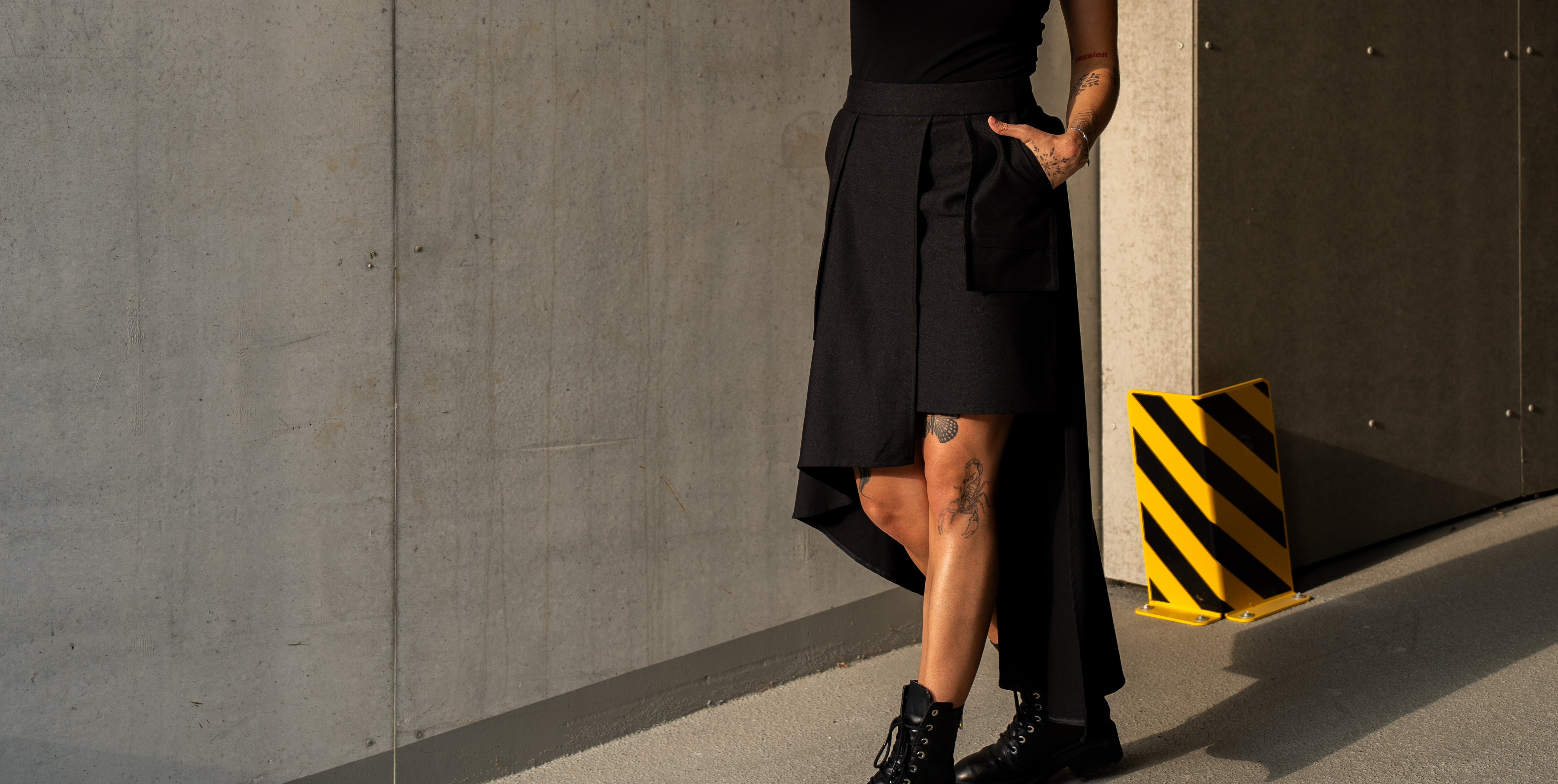 Person wearing a black skirt standing against a concrete wall with a caution sign.