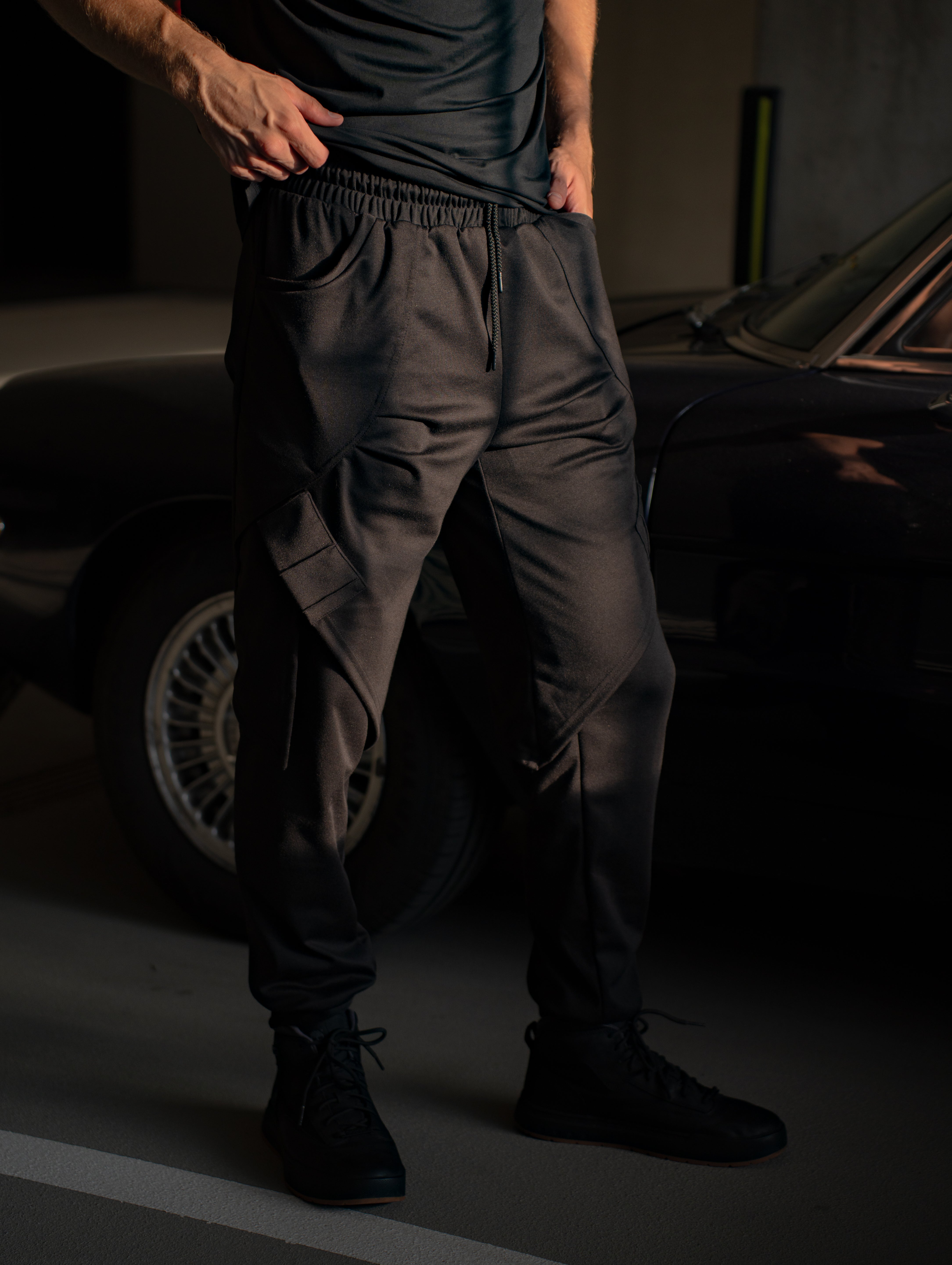 Person close up of dark trending techwear clothing wearing dark pants standing in a dimly lit garage with a car in the background