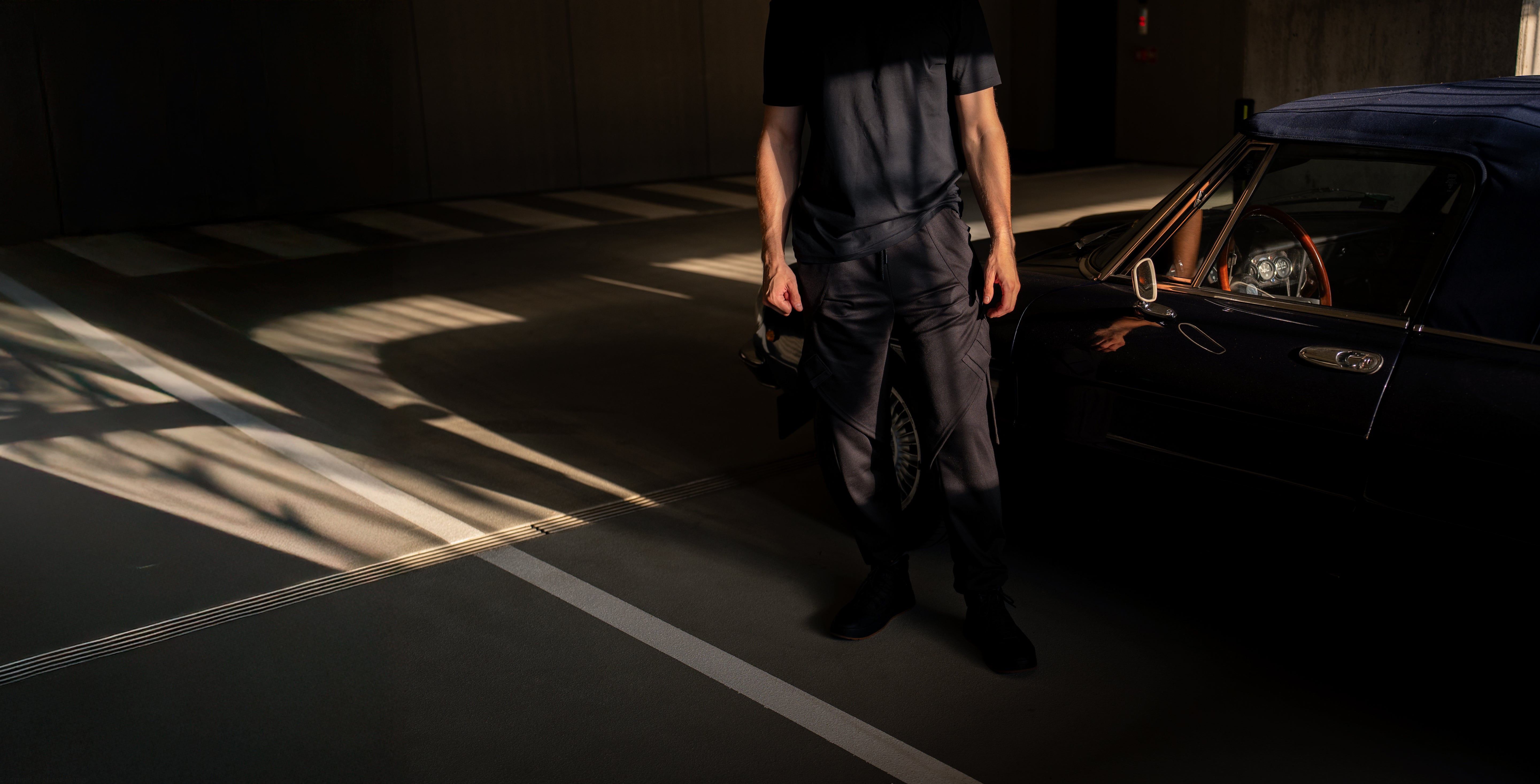 Person standing next to a classic car in a dimly lit garage wearing all black new urban streetwear
